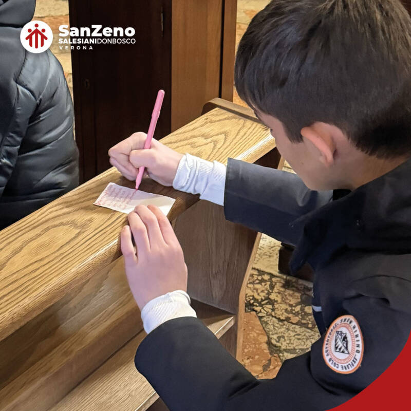 Un ragazzo sta scrivendo su un foglio di carta con una penna rosa. È seduto su una panca all'interno di una chiesa. Si tratta di studenti e studentesse dell'Istituto Salesiano San Zeno di Verona.