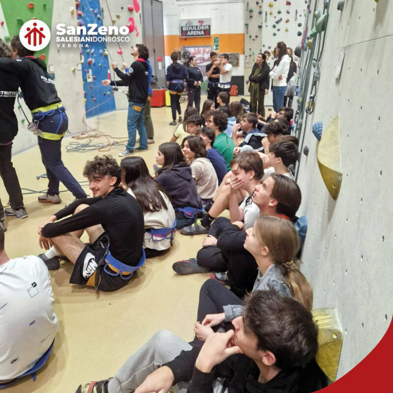 Un gruppo di giovani è seduto per terra e guarda i compagni arrampicarsi su una parete di roccia. Si tratta di studenti e studentesse dell'Istituto Salesiano San Zeno di Verona.